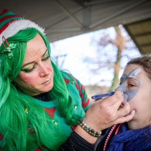 Maquilleuse déguisée en lutin de Noël réalisant un maquillage artistique sur le visage d’un enfant lors d’un événement festif.
