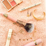 Flatlay délicat regroupant bijoux dorés et produits de maquillage favoris sur une serviette rose, illustrant l’univers intime et sensoriel de Léa.