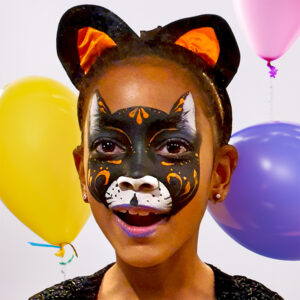Enfant maquillée en chat noir avec des oreilles assorties et des ballons colorés en arrière-plan lors d’un anniversaire.