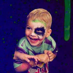 Jeune garçon maquillé façon Joker, riant pendant un événement festif, fond artistique vert et violet.