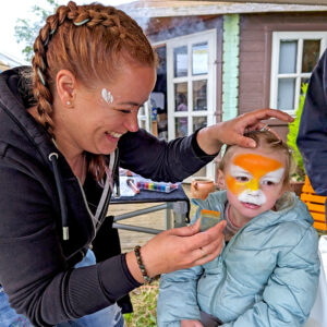Maquilleuse souriante en pleine réalisation d’un maquillage de tigre sur le visage d’une petite fille pendant une animation.
