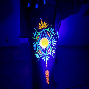 Maquillage fluo UV coloré sur jambe sombre, motifs tribaux lumineux sous lumière noire.