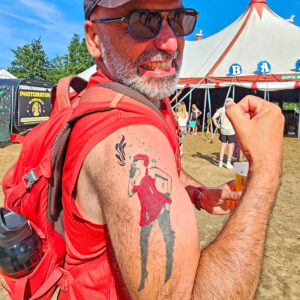 Homme souriant en festival arborant un tatouage éphémère représentant une femme en robe rouge.