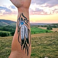 Maquillage artistique de plumes et motif tribal peint sur l’avant-bras, photographié en pleine nature au coucher du soleil.